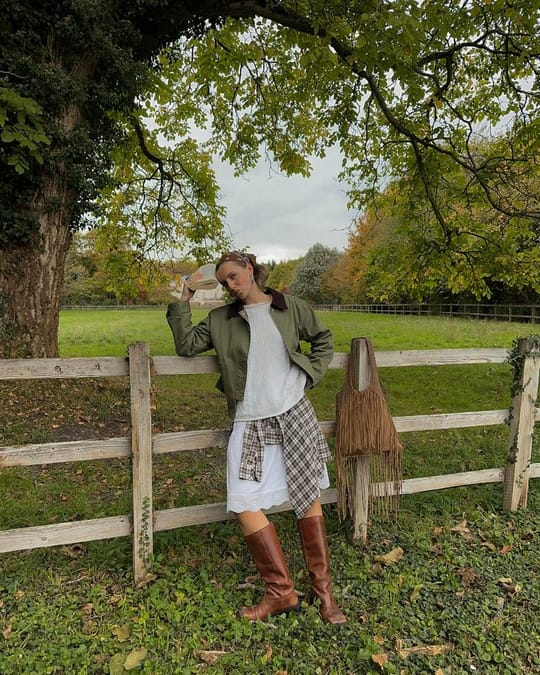 Woman in white skirt, brown boots and green barn jacket leaning on a fence